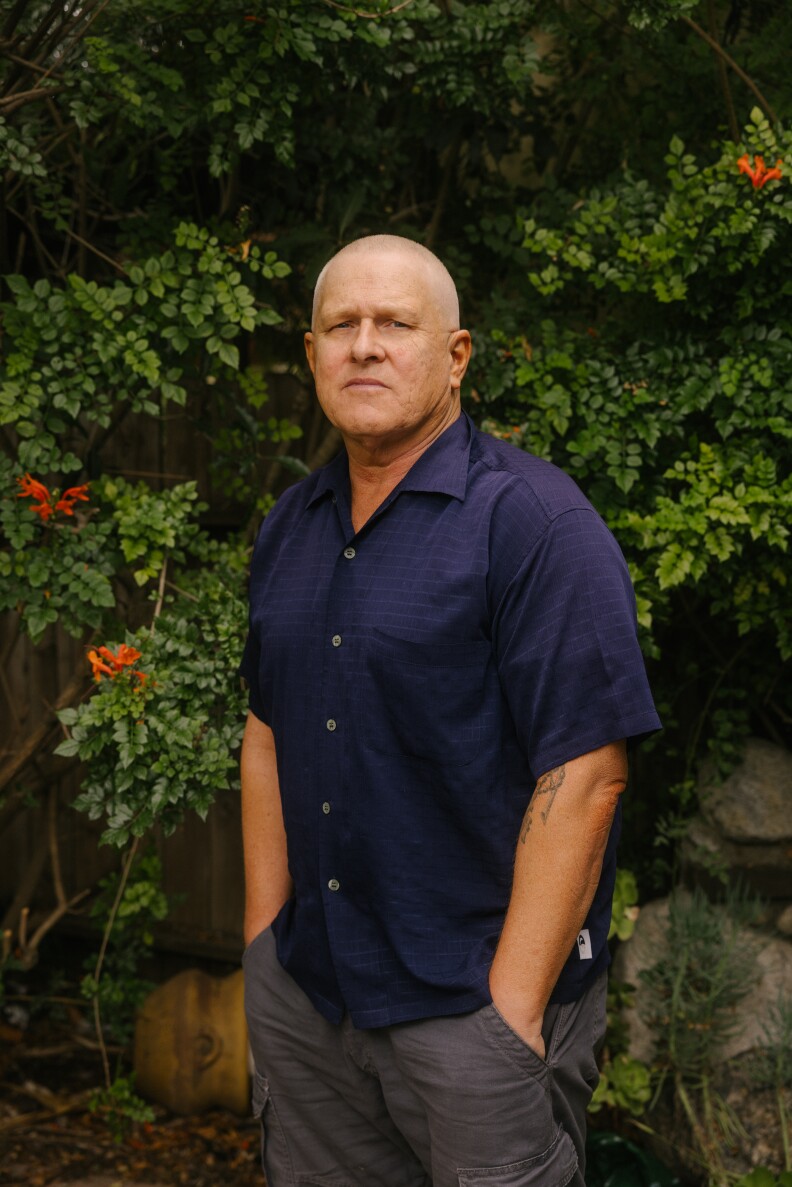 A bald man with light-brown skin tone, wearing a short-sleeved blue shirt and gray slacks, stands in front of a green hedge