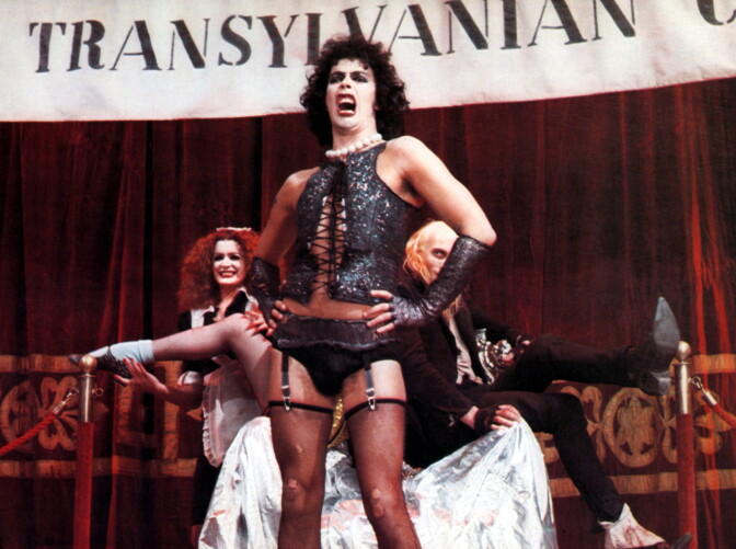 Tim Curry as Dr. Frank-N-Furter in The Rocky Horror Picture Show. He wears curly black hair, a white pearl necklace, a sparkly black vest, black panties, black gloves, and black thigh-high stockings.
