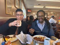 A light brown man holding a coffee cup on the left and a Black man holding a fork on the right, eating at a diner in Inglewood.