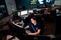 A woman with dark hair in a ponytail and wearing a blue t-shirt sits at a desk in front of several computer screens answering 911 calls.