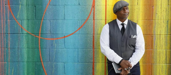 A black man is pictured on the right side of the photo against a wall painted in bright colors. He looks off to the side. He is dressed in a button-up vest, tie and white dress shirt, and a newsboy-style cap. 