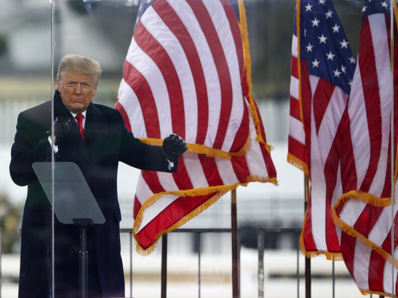 Donald Trump wears a dark trench coat with red tie while standing at a lectern.