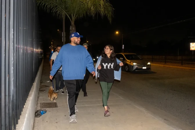A group of people walk on a sidewalk past a metal fence and car driving by on the street at night.