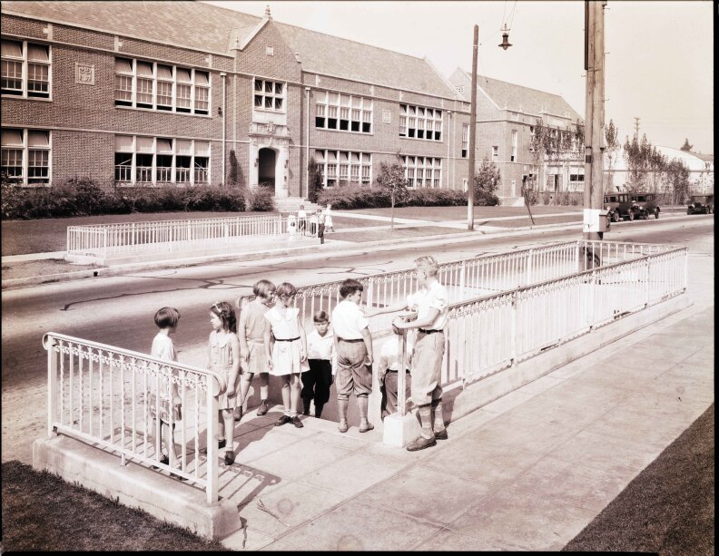 A black and white image shows children at the top of the stairs of a pedestrian tunnel. A large school building is seen across the street in the background.