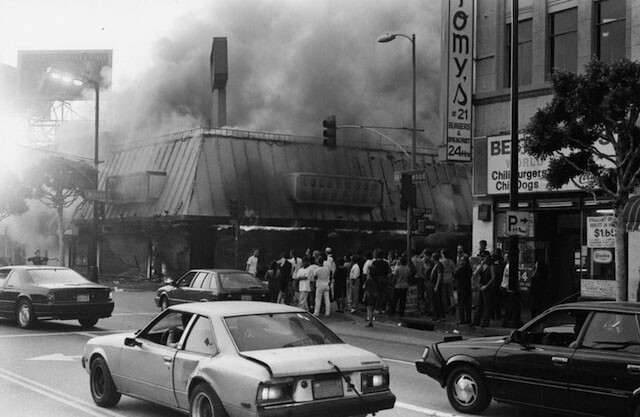 A black-and-white photo of a Los Angeles street, cars are crossing in the foreground, in the background a building is burning and a large crowd is gathered in front of it.