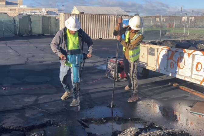 Two persons use construction tools. They wear white hard hats.