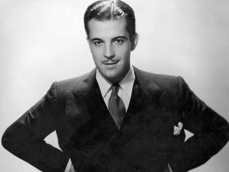 A man in a dark suit and tie, with a pocket square, has a mustache and slicked back hair.