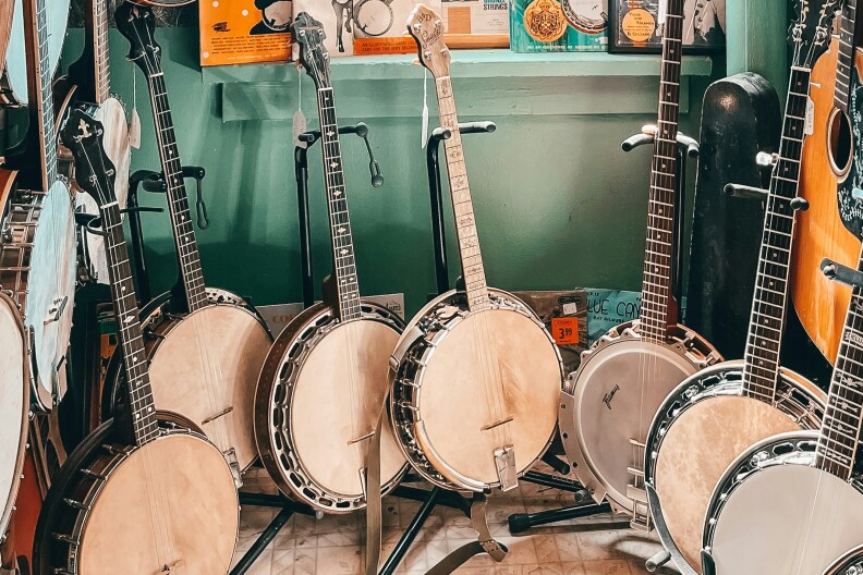Image of a room full of Banjo guitars and a few different types too. All presented in a small greenish teal room. Some are hanging while others are on the floor on guitar stands.