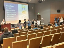A lecture-hall style room with dozens of chairs arranged in a semi-circle facing the stage, podium, and screen at the front. The chairs are mostly empty as small groups of people talk to each other in various corners of the space.