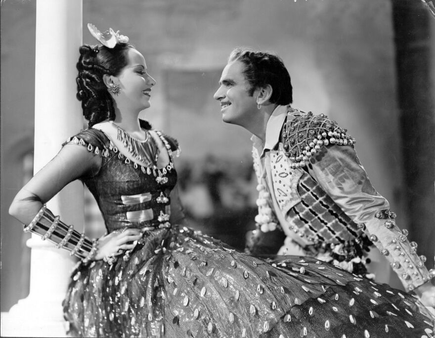 Merle Oberon wears an extravagant dress which appears to have Eastern flair, while a man also in an extravagant outfit gives her an adoring look. The image is in black and white.