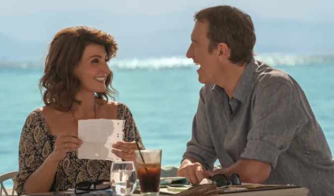 Nia Vardalos and John Corbett sit a table oceanside, bright blue waves crashing  behind them. Vardalos looks at Corbett smiling. She is holding a piece of paper that seems to be a page ripped from a notebook. Corbett looks at her, his mouth open as if laughing. 