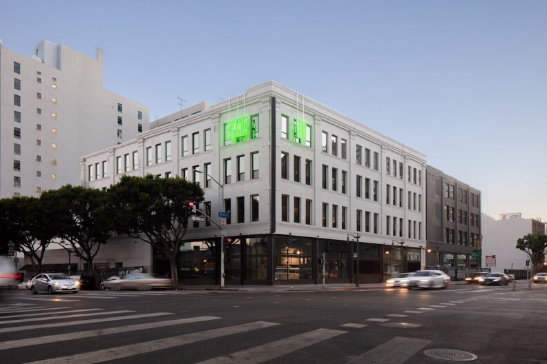a gray building on a corner has two windows lit up in neon green as cars drive past