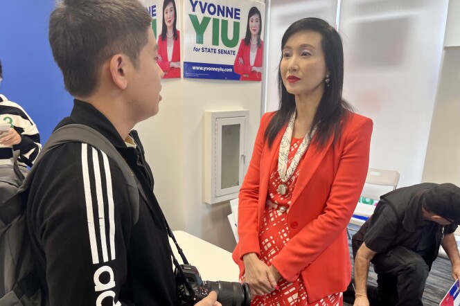 A 52-year-old Asian American woman in a bright red blazer and dress looks at a young Asian American man whose mostly faced away from the camera. A campaign sign attached to a pillar behind her. It reads "Yvonne Yiu for State Senate."