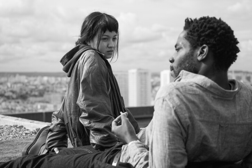 A woman and man sit atop a roof overlooking the city of Paris. The woman is staring at the man, who is looking off into the distance.