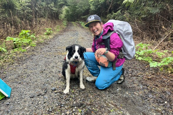 A tired border collie sitting next to a woman in hiking gear smiling big at the camera.