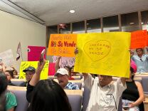 A group of residents it in blue chairs in Santa Ana City Hall holding brightly colored signs that read "Santa Ana no se vende" or "Santa Ana is not for sale," and "Santa Ana no se vende, se cuida" or "Santa Ana is not to sell, its to take care of." 