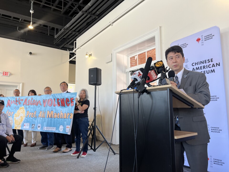 A Japanese American man speaks at a podium as people to his side hold a blue banner that says "Stop Anti-Asian Violence."