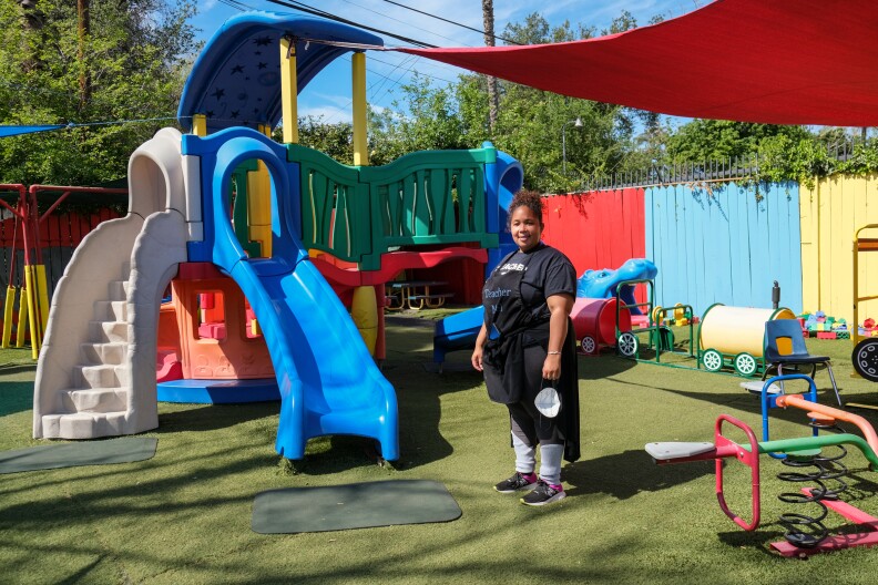 A woman stands in the middle of a preschool playground next to a bright blue slide.