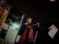 A woman with glasses and a red scarf speaks into a microphone and holds a clipboard with a paper attached to it.
