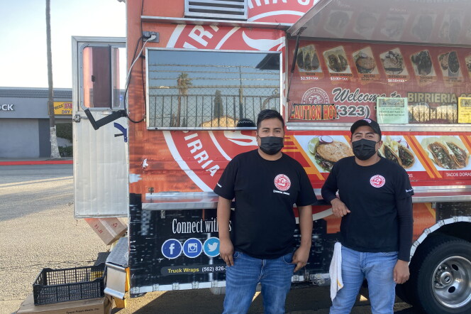 two men in black shirts and blue jeans, both wearing black face masks, stand near the rear of a food truck.