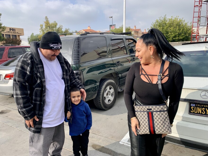 Three people, two adults and a child, stand in a parking lot. A man has his arm around the child.