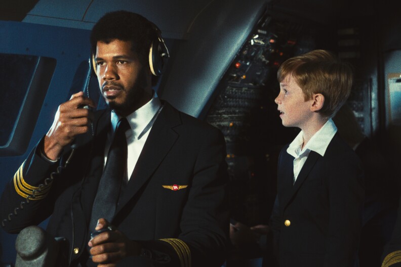 Solomon Hughes, playing Kareem Abdul-Jabbar, in a navy pilot's jacket on the left and holding an intercom mic. On the right is a small redhead child in a suit jacket. They are on an airplane set.