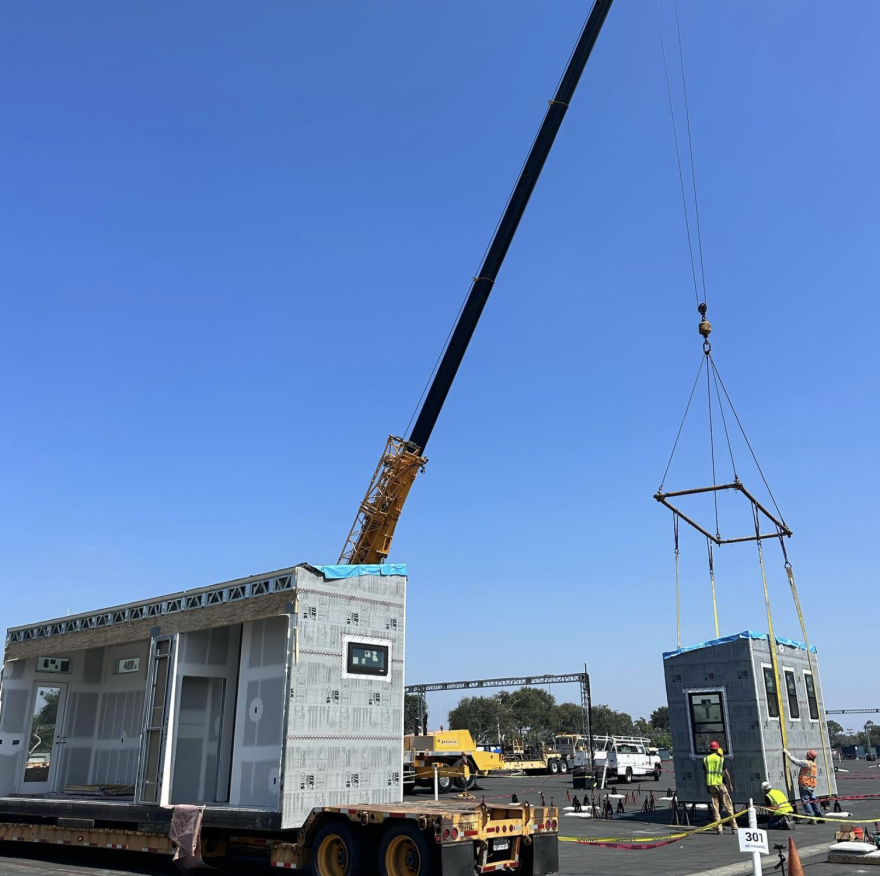Students stand below a long crane machine, cradling the small portion of a house in pieces. The main portion of the house is at the front and to the left of them. They are all wearing hard hats and safety vests.