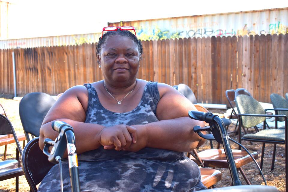 A woman with dark-tone skin wears a sleeveless blue tie-dyed dress sits in a chair, looking at the camera. She's sitting among dozens of empty folding chairs in the garden