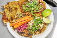Overhead view of a white paper plate holding three tacos on crisp, griddled tortillas: one folded with melted cheese along the edges, one topped with mushrooms, refried beans, red onion, and cilantro, and one topped with grilled chicken, onion, and cilantro. Carrot sticks, pickled red onions, and lime wedges sit alongside.
