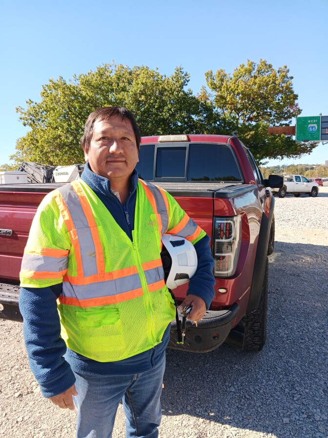 A man with medium skin tone, wearing a yellow safety vest and holding a white safety helmet, stands behind a red pick up truck on a gravel road.