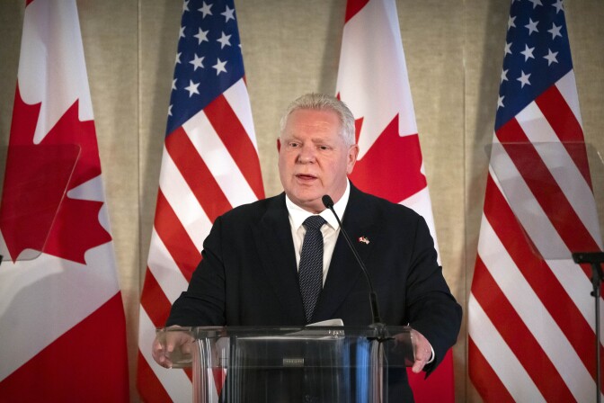 A man in a dark suit speaks at a lectern in front of U.S. and Canadian flags