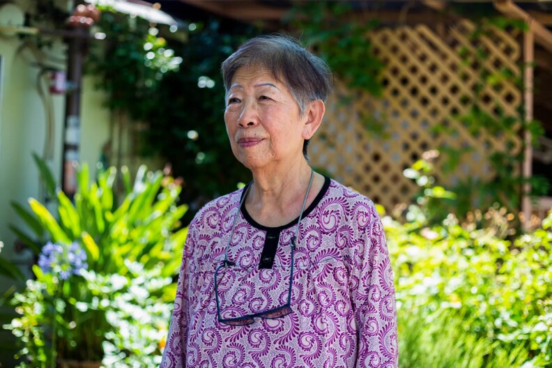 An older woman wearing a purple blouse poses for a photo in front of some plants.
