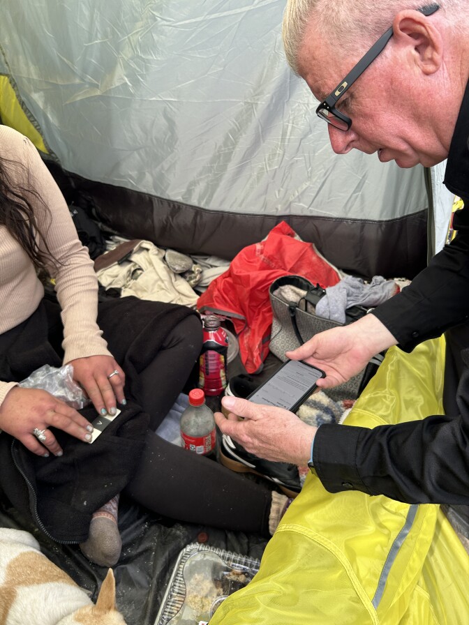 The inside of a tent showing, on the left side, a woman's hands with her fingers pressed on a mobile EKG monitor, and on the right, a man's face in profile. He's holding a cell phone monitoring the device. On the floor of the tent are soda bottles and clothes, and you can part of a small white and tan dog. 