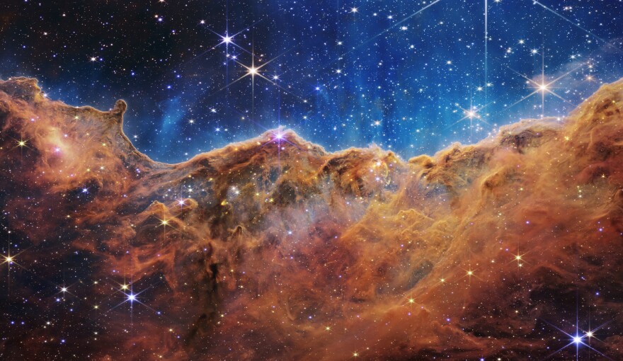 A very clear image of outer space shows a gaseous, brown and rust colored landscape of mountains and valleys speckled with glittering stars against a black sky.