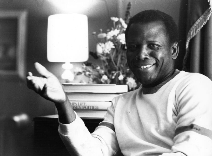 Sidney Poitier in a shite long sleeve shirt is smiling as he gestures with his right hand.