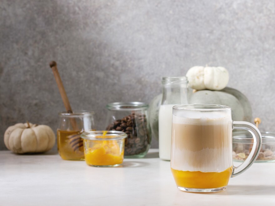 A glass mug contains an orange-tinted liquid. A small pumpkin, jar of honey and other small containers are seen in the background.
