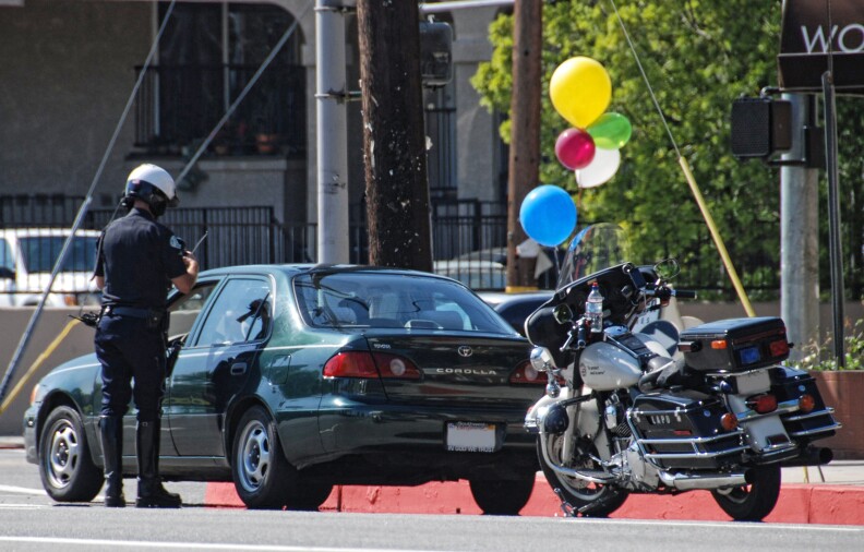 A police officer stands next to a dark green Toyota Camry, writing the driver a traffic ticket.