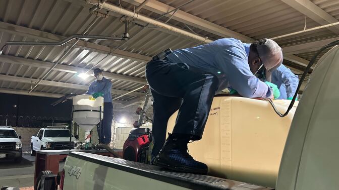 A wide shot of two vector district staff members working with large containers at night. One is to the left at the back working with the big spout. The other is to the right on the truck bed crouching down to work with tools on the wter tank.