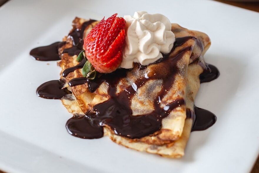 An image of a crepe sits on a white plate. The crepe is a light brown color and is covered with a dark brown chocolate sauce, a piped dollop of whipped cream and half a strawberry