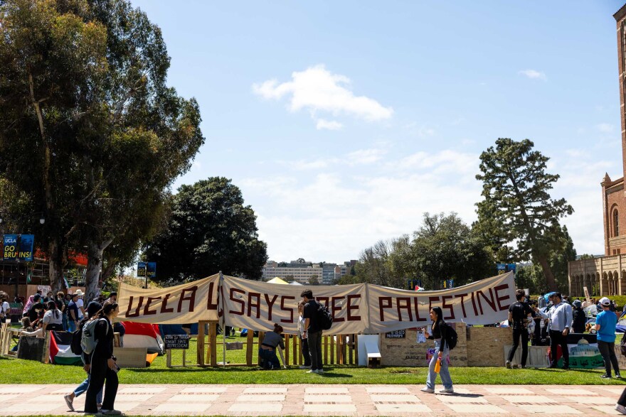 A large banner on a lawn next to a concrete walkway that reads "UCLA SAY FREE PALESTINE." Various people stand around the sign on the lawn.