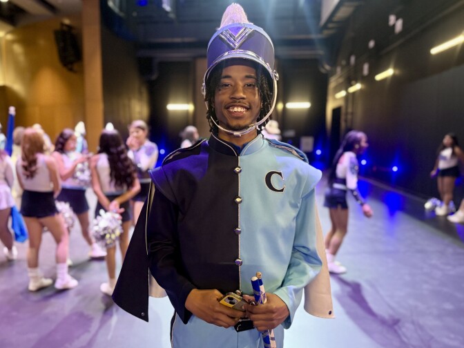 A student with dark brown skin tone in a blue marching band uniform smiles for the camera. Behind him are students in cheerleading attire on a stage.