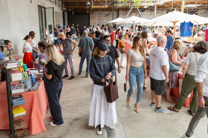 Several dozen people walk across a courtyard buying books. A woman in the foreground wears a blue hat, blue sweatshirt, a white skirt, and carries a brown bag. She is putting something into the bag. People can be seen walking and in conversation behind her.