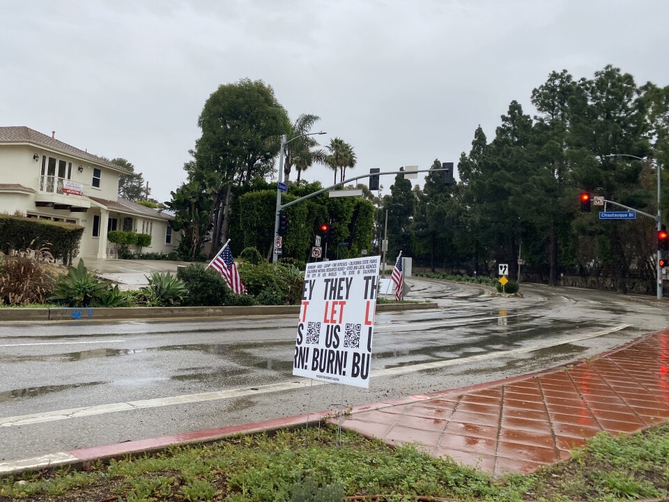 A small sign that reads "They let us burn!" stands on a patch of grass near a street intersection across from a home and some trees.