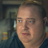 Brendan Fraser sits right of frame, only his shoulders up are visible. Fraser wears a gray shirt, his hair thin and stringy on top of his head. He looks off to the left. His expression is sad, his eyes slightly hooded under faintly furrowed brows. His lips are slightly parted. Behind him seems to be a kitchen with brown cabinets and clutter on the countertops. 