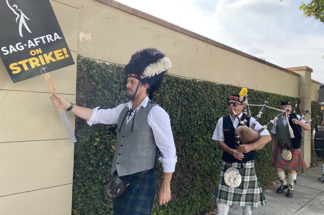 A bagpipe band marches in front of Warner Bros. studios in Burbank. They are in front of a beige wall with green ivy on it. The band wears kilts and traditional Scotiysh garb