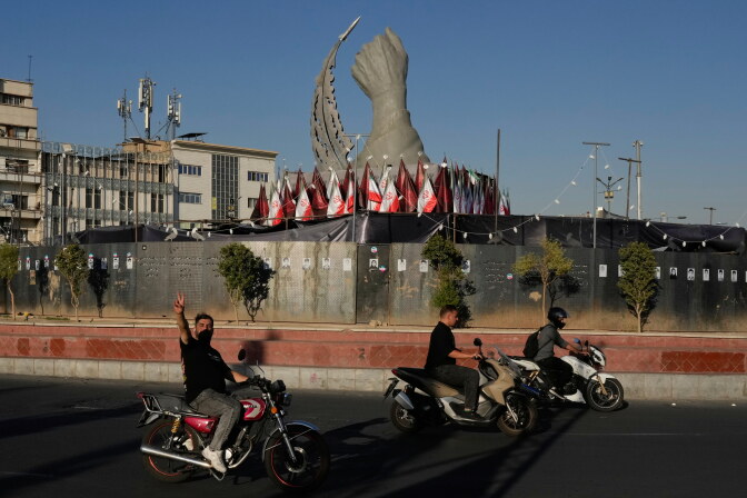 Three men ride their scooters around a town square.