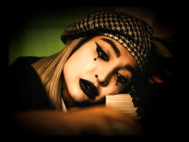 A woman in white face paint with black lips and exaggerated black mime eyes rests her head on the keys of a piano while looking sad.
