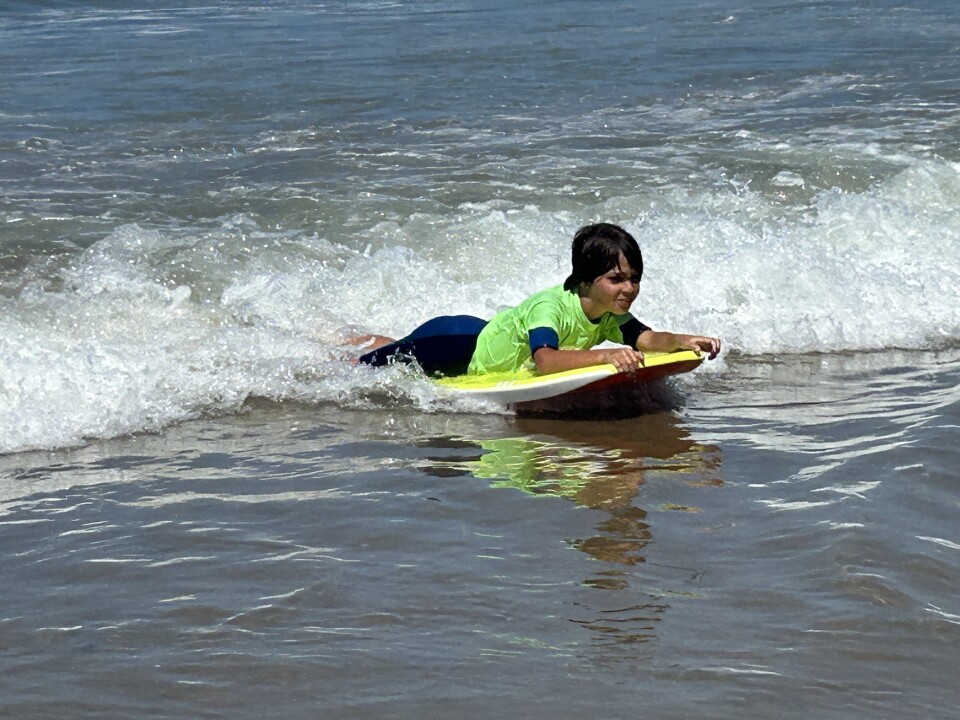 A boy in a neon green shirt over a short wetsuit rides a boogie board in on a small wave. 