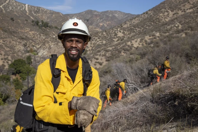 A male-presenting person clad in a bright yellow jacket, large, dirty gloves, a white helmet, and black backpack stands against a mountainous backdrop. The hills are beige and brown, with a lot of dry vegetation and gray sticks. There is scarce greenery to the left of him. Several people in yellow and orange clothing and helmets walk up the hill behind him carrying gear.