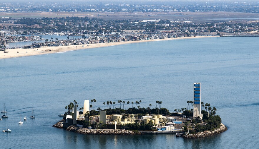 Aerial photo of a small island off a coastline populated with various buildings. The island is ringed with palm trees and four tall structures. In the middle of the island are several round buildings. To the left of the island, six small boats are pictured, in a cluster